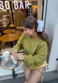 Pull en tricot vert avec des manches larges, deux tasses de café sur une table ronde, et un sac à main matelassé jaune avec une bandoulière en chaîne dorée.