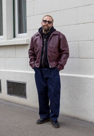 Chaqueta de cuero marrón con dos bolsillos frontales, sobre una sudadera negra, combinada con pantalones de mezclilla azules anchos y zapatos negros, de pie contra una pared.