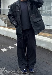 Crna vodootporna jakna s dva džepa na prsima, uparen s širokim crnim hlačama i plavim patikama s uzorkom, stoji na popločenoj površini.