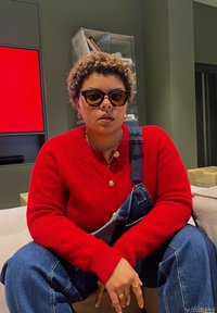 Κόκκινο ριγέ πουλόβερ, μπλε τζιν σαλοπέτα, στρογγυλά γυαλιά ηλίου από καβούκι χελώνας. Καθισμένος/η σε ένα ουδέτερο φόντο με μια τηλεόραση που εμφανίζει μια κόκκινη οθόνη.