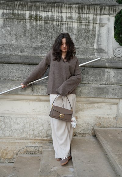 Suéter marrón de punto texturizado con escote en V, combinado con pantalones amplios claros. Lleva un bolso marrón con un cierre circular y un charm.