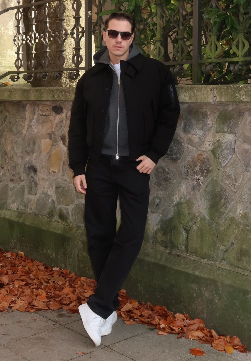 Zwarte bomberjas over een grijze sweater, gecombineerd met zwarte jeans en witte sneakers. Tegen een stenen muur met herfstbladeren als achtergrond.