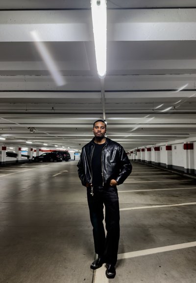 Hombre con chaqueta de cuero negra, pantalones negros y botas, de pie con las manos en los bolsillos en un estacionamiento poco iluminado y mayormente vacío.