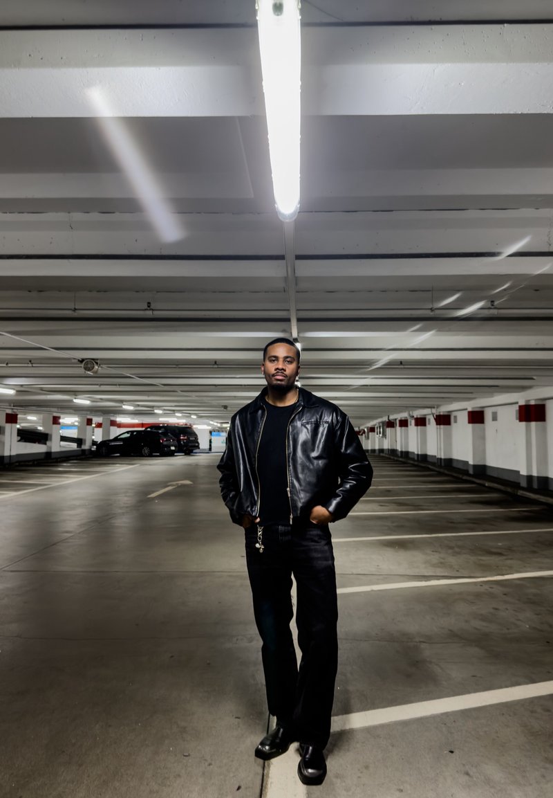 Homme en veste en cuir noire, pantalon noir et bottes, debout les mains dans les poches dans un parking peu éclairé et presque vide.
