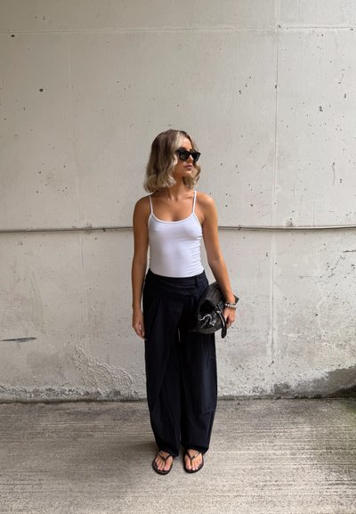Top blanco sin mangas, pantalones de pierna ancha plisados en negro, bolso negro, chanclas y gafas de sol; de pie contra una pared gris texturizada.