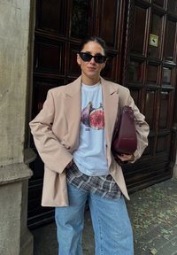 Sieviete valkā melnas saulesbrilles, bēšu pārāk lielu žaketi, baltu T-kreklu ar augļu grafiku, dzeltenu kreklu, kas sasiets ap vidukli, gaišus zilo džinsus un tur sarkanā burgundijas krāsā rokas soma.