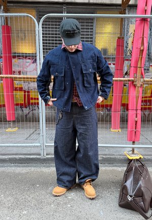 Persona con chaqueta de mezclilla oscura y jeans anchos, botas color topo, gorra negra, de pie en la calle cerca de una cerca metálica y una bolsa marrón en el suelo.