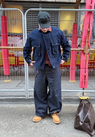 Persona con chaqueta de mezclilla oscura y jeans anchos, botas color topo, gorra negra, de pie en la calle cerca de una cerca metálica y una bolsa marrón en el suelo.