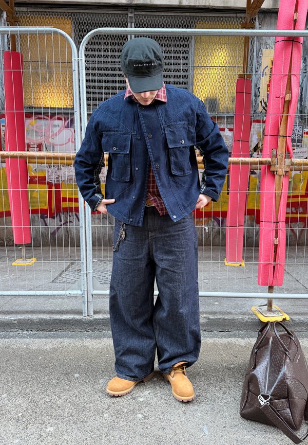 Sötét farmer dzsekit és széles szabású farmert viselő személy, világosbarna csizmában, fekete sapkával, az utcán áll, egy fémkerítés mellett, a földön barna táska van.