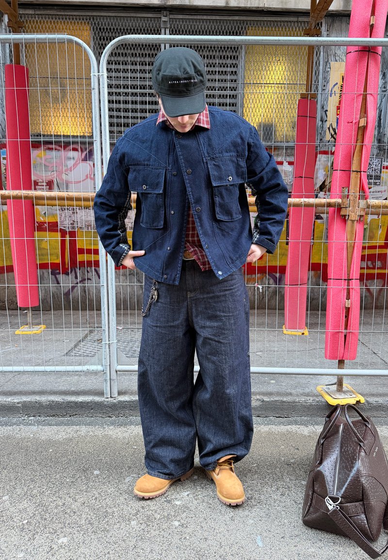 Persona con chaqueta de mezclilla oscura y jeans anchos, botas color topo, gorra negra, de pie en la calle cerca de una cerca metálica y una bolsa marrón en el suelo.