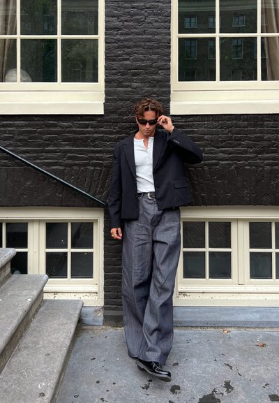 Blazer negro sobre una camiseta henley gris, combinado con pantalones anchos de rayas grises y zapatos negros, frente a una pared texturizada negra.