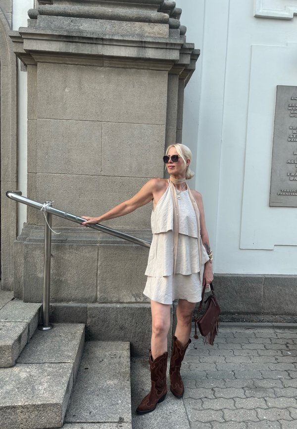 Žena v béžových šatách bez rukávov, hnedých kovbojských topánkach a slnečných okuliaroch stojí vedľa kamenných schodov, drží kovové zábradlie a hnedú kabelku.