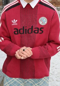 Punane kootud kampsun musta "adidas" kirjaga, valgete triipudega varrukatel ja ümmarguse logoga. Mudeli käed kannavad hõbedast sõrmust.