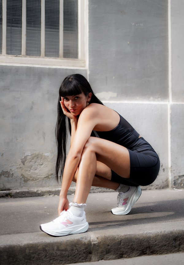 Jeune femme aux longs cheveux noirs vêtue d'une tenue de sport noire, qui lace des baskets blanches en étant accroupie sur un trottoir, près d'un mur en béton gris.