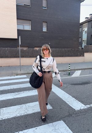 Blusa blanca de manga larga con estampados gráficos en negro, pantalones marrones de cintura alta y un gran bolso de hombro negro acolchado. Cruzando una calle.