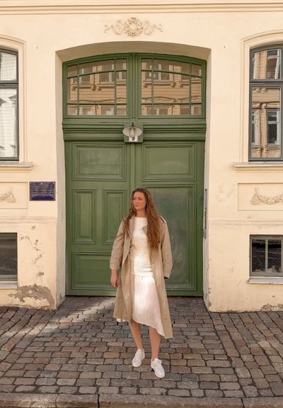 Mujer con cabello largo, con gabardina beige y vestido blanco, de pie en una calle empedrada frente a grandes puertas verdes y una fachada de edificio color crema.