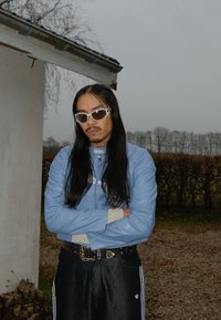 Veste en cuir bleu clair avec accents blancs, associée à un pantalon large noir avec des bandes blanches. Accessoirisée de lunettes de soleil blanches et d'une ceinture cloutée.