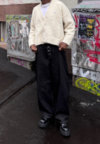 Krém színű, puha kardigán V-nyakú kivitelben, alatt fehér póló, hozzá magas derekú, fekete, bő szárú farmer és masszív fekete csizma.