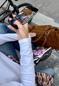 Pametan telefon u ruci, prikazuje smeđi torbu i osobu koja nosi bijelu majicu dugih rukava, plave traperice i tenisice s uzorkom.