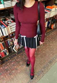 Top de manga larga de canalé burdeos combinado con una falda negra con pliegues que presenta detalles en cinta blanca y medias rosas, terminado con zapatos negros de charol.