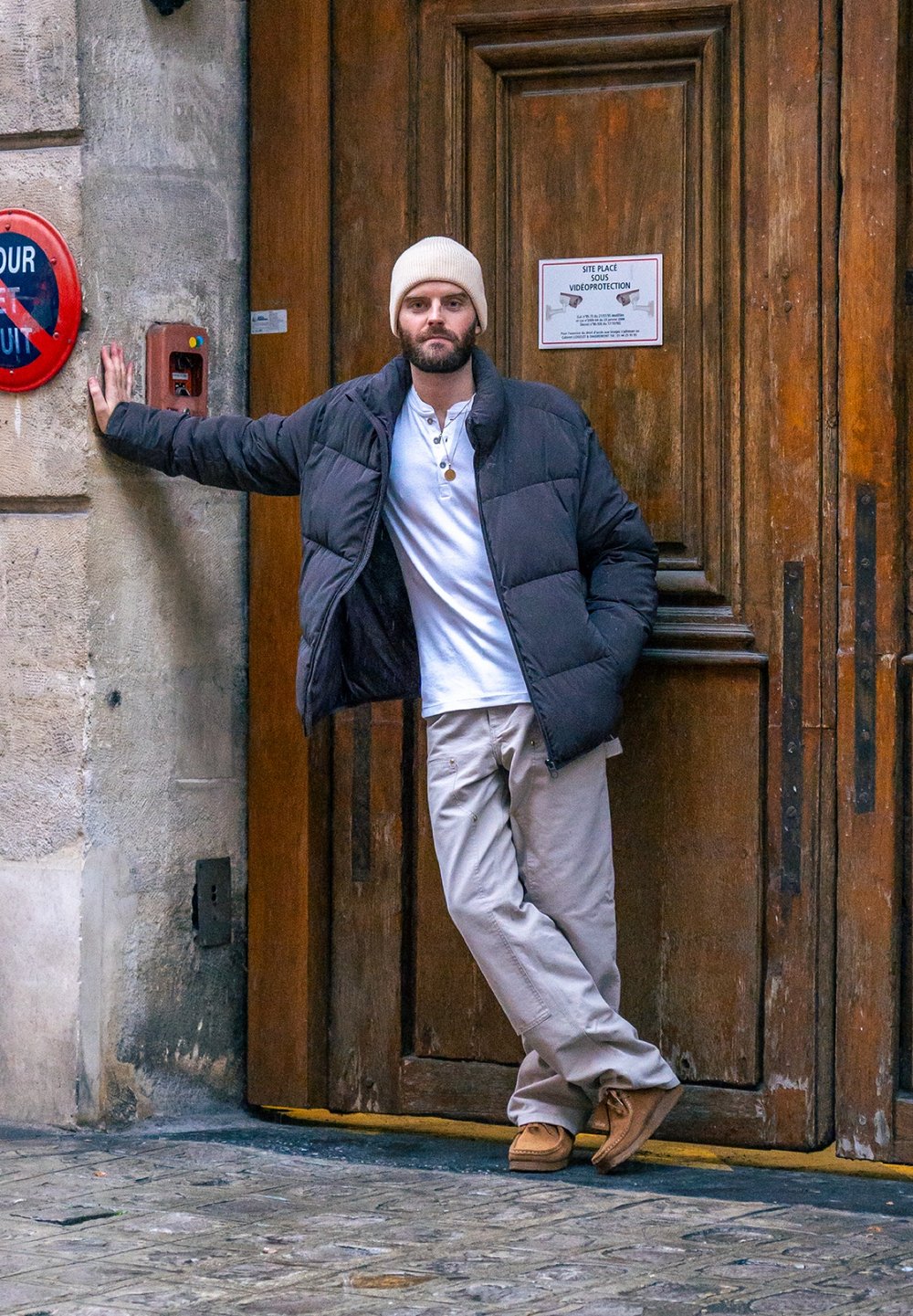 Doudoune noire, chemise blanche à boutons, pantalon beige et chaussures marron clair. Un homme s'appuie contre une porte en bois avec un panneau rouge.