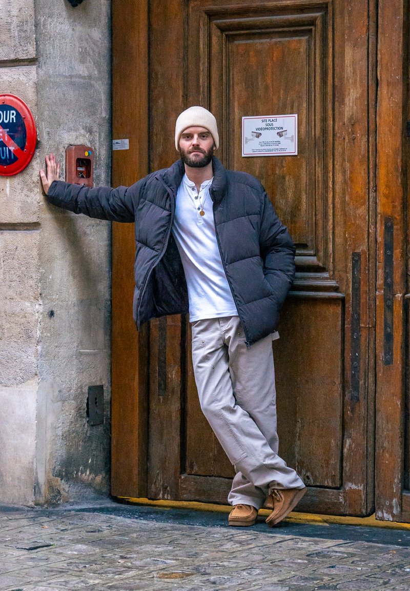 Doudoune noire, chemise blanche à boutons, pantalon beige et chaussures marron clair. Un homme s'appuie contre une porte en bois avec un panneau rouge.