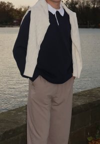 Marineblaues Langarmshirt, kombiniert mit einer cremefarbenen Zopfmuster-Strickweste und beigefarbenen Hosen. Das Outfit zeichnet sich durch einen klassischen Kragen und eine lockere Passform aus.