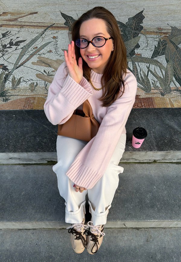 Mujer joven con gafas, suéter rosa, pantalones blancos y zapatillas beige saludando mientras está sentada en escalones de piedra junto a una taza de café rosa.