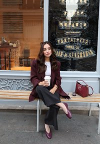 Blazer color borgogna, top bianco e pantaloni neri a gamba larga. Tiene in mano una tazza di caffè; borsa color bordeaux, scarpe abbinate e panchina di legno con texture.