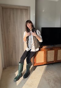 Femme assise sur une armoire en bois près de la porte, ajustant un gilet vert foncé par-dessus une chemise à carreaux beige, portant un pantalon noir et des bottes de pluie vertes.