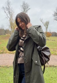 Woman adjusting the collar of a green coat, wearing a patterned sweater, carrying a dark brown bag, standing outdoors in an autumn park.