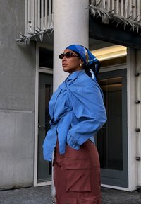 Camicia azzurra oversize con nodo sul davanti, gonna cargo marrone con tasche e foulard a fantasia blu. Occhiali da sole scuri.