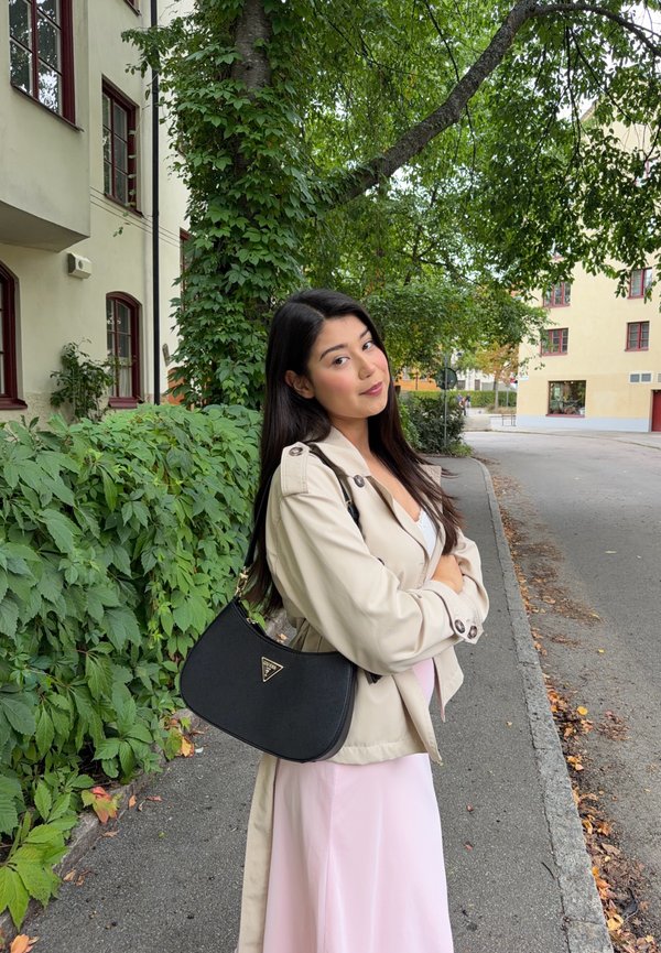 Une femme portant une veste beige et une robe rose pâle se tient sur un trottoir urbain couvert de feuilles, un sac noir en bandoulière.