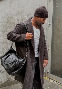 Black leather bag with a textured finish, carried by a person in a dark brown, textured overcoat, in front of a concrete background.