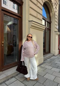 Rævet rosa trøje med striber, hvide bukser med vide ben, brune ruskindsstøvler og en mørkebrun håndtaske. Står op ad en bygning med store vinduer.