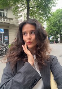 Une personne portant un blazer gris à rayures avec de grands revers et un haut blanc, touchant son visage, dans un environnement urbain en plein air.