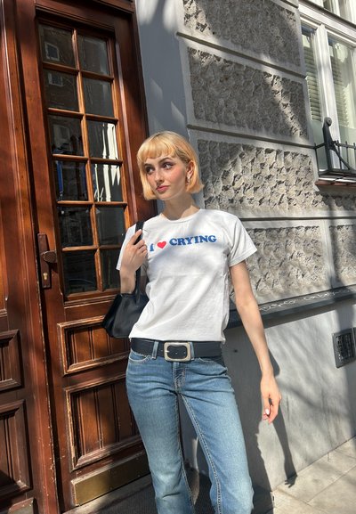 Camiseta blanca de algodón con el texto azul "I ❤️ CRYING", combinada con jeans de un azul claro. Un cinturón negro y un pequeño bolso negro complementan el conjunto.