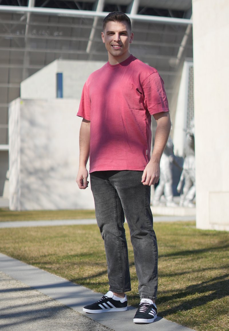 Jeune homme debout en plein air sur l'herbe, portant un t-shirt rose, un jean noir et des baskets Adidas noires, souriant à la caméra.