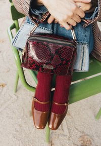 Des mains tenant un sac à main en cuir de serpent rouge et noir reposant sur une jupe en jean, avec des chaussettes rouges et des chaussures Mary Jane marron, assis sur une chaise verte.