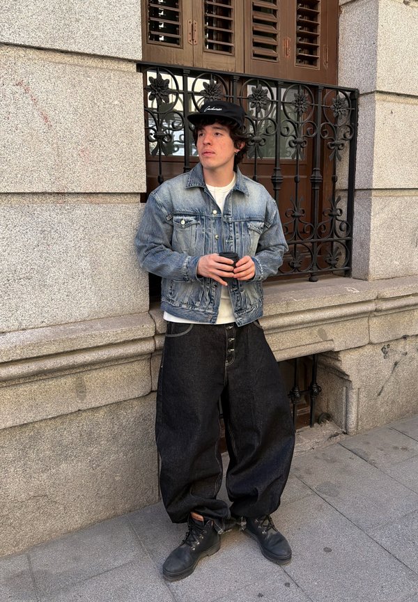 Ung mand i cowboyjakke og løstsiddende sorte bukser, der holder en smartphone og står foran en stenmuret bygning med et smedejernsvinduesgitter.