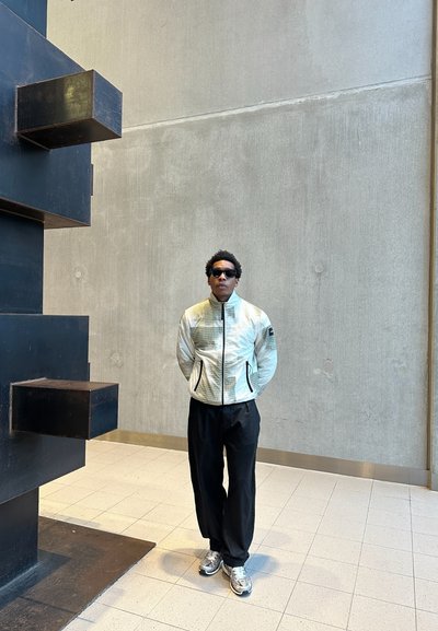 Chaqueta texturizada de color claro con cremallera en el frente, pantalones negros y zapatillas metálicas. Presentada frente a una pared gris y una estructura abstracta negra.