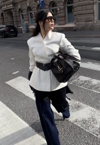 Weißer, strukturierter Blazer mit schwarzem Gürtel, kombiniert mit dunklen Denim-Hosen mit Spitzenakzenten. Accessoires sind große Sonnenbrillen und eine Lederhandtasche.