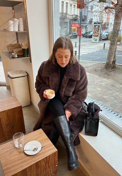 Abrigo de piel sintética marrón, botas negras hasta la rodilla, sosteniendo una taza blanca. Mesas de madera y un vaso de agua con un plato blanco y una cuchara.