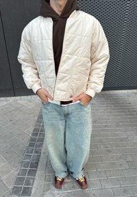 Hellbeige gestepptes Bomberjacke mit Reißverschluss, getragen über einem braunen Hoodie. Kombiniert mit lockeren hellblauen Jeans und braunen Turnschuhen.