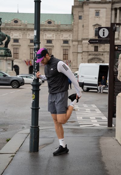 Atleta con una chaqueta sin mangas negra, camiseta de manga larga blanca, pantalones cortos grises y zapatillas negras, estirándose al lado de una farola en un entorno urbano.