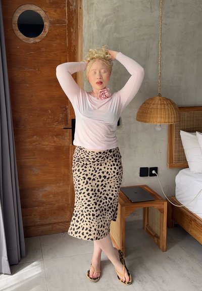 Mujer con el cabello rubio trenzado y peinado hacia arriba, vistiendo una camiseta rosa de manga larga y una falda con estampado de leopardo, de pie con los ojos cerrados en un dormitorio moderno.