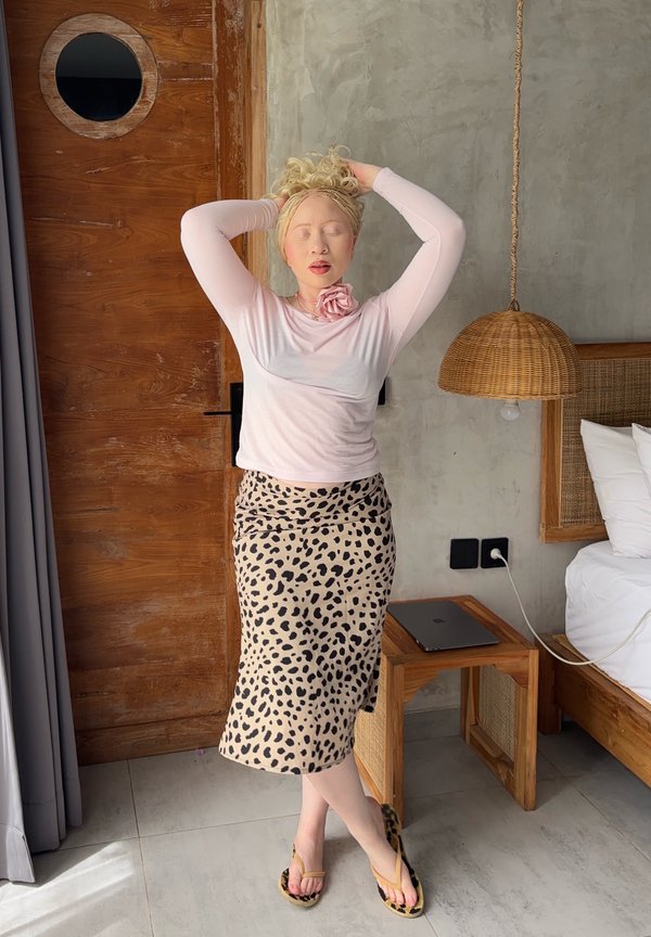 Žena s vrkočmi blond vlasov upravených hore, oblečená v ružovom top s dlhým rukávom a sukni s leopardím vzorom, stojaca so zatvorenými očami v modernej spálni.