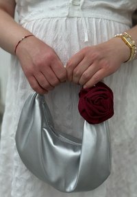 Händer som håller en silverhandväska med en röd tygros fäst, mot en vit texturerad klänningsbakgrund.