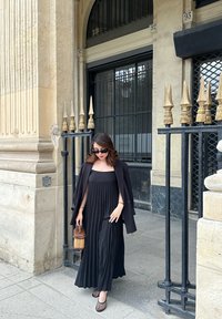 Schwarzes, plissiertes Maxikleid mit trägerlosem Design, kombiniert mit einem schwarzen Blazer, der eine kleine braune geflochtene Tasche hält, vor dem Eingang eines Gebäudes.