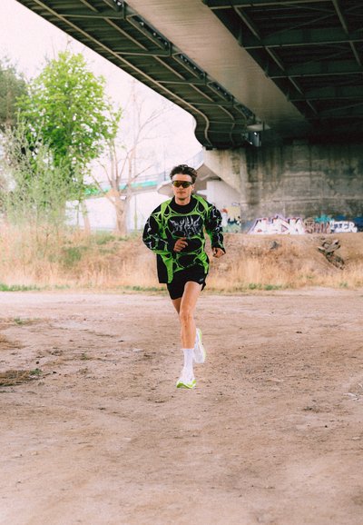 Hombre con ropa deportiva de color verde neón y negro y gafas de sol corre bajo un puente de hormigón con grafitis en la pared detrás.